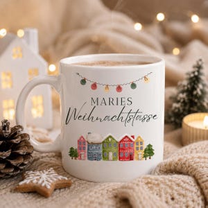 Peut inclure: Un mug en céramique blanche orné d'une guirlande de lumières colorées, du texte "MARIES Weihnachtstasse" en noir, et une rangée de petites maisons illustrées colorées avec des arbres verts à la base, rempli d'une boisson brune chaude.