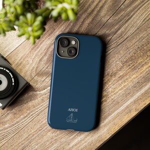 Puede incluir: Una funda de smartphone azul oscuro con una ilustraci&oacute;n de l&iacute;neas blancas de un velero y el texto AHOI impreso en blanco. La funda tiene un recorte para la c&aacute;mara con un borde gris para un dispositivo con sistema de doble lente.