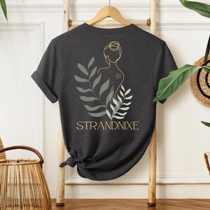 May include: Dark gray t-shirt with a line drawing of a woman with a star on her back, surrounded by green leaves. The text "STRANDNIXE" is printed below the drawing.