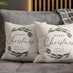 Könnte beinhalten: Zwei weiße Kissen mit dem Schriftzug "MERRY Christmas" in eleganter Schrift, umgeben von grünen Blättern. Der Text "FAMILIE SCHMIDT" steht darunter. Die Kissen liegen auf einem grauen Sofa.
