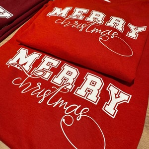 Peut inclure: T-shirts rouges avec l'inscription "MERRY christmas" en lettres majuscules blanches et en écriture cursive. Un t-shirt est plié sur un autre. Un t-shirt bordeaux est visible en arrière-plan.