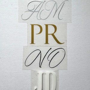 Peut inclure: Quatre lettres en vinyle, chacune avec une police et une couleur différentes. La lettre du haut est argentée et dit "HM", la deuxième lettre est dorée et dit "PR", la troisième lettre est noire et dit "VO", et la lettre du bas est blanche et dit "JD".