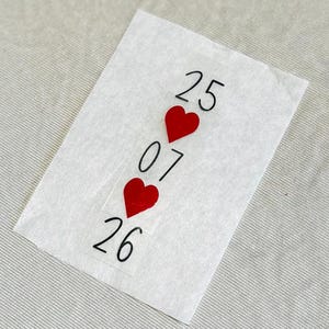 Puede incluir: Un papel blanco con los números 25, 07 y 26 en negro, separados por formas de corazón rojo. El papel es ligeramente transparente y descansa sobre una superficie gris claro.