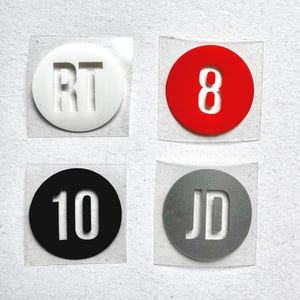 May include: Four small, square stickers with circular designs. One white sticker has the letters "RT", a red sticker has the number "8", a black sticker has the number "10", and a grey sticker has the letters "JD".