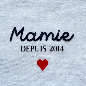 Peut inclure: Sac fourre-tout blanc cassé avec le mot "Mamie" en cursive noire, "DEPUIS 2014" en police sans-serif et un cœur rouge. Le sac est fait d'un tissu texturé.