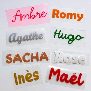 Può includere: Una collezione di decalcomanie con nomi in vari colori e caratteri. I nomi includono "Ambre", "Romy", "Agathe", "Hugo", "Sacha", "Rose", "Inès" e "Maël". Le decalcomanie sono su supporto trasparente, con nomi in rosso, arancione, verde, marrone, bianco e oro.