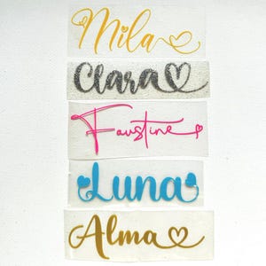 Op de afbeelding: Een verzameling gepersonaliseerde naamstickers in verschillende kleuren. De namen "Mila" in goud, "Clara" in zilveren glitter, "Faustine" in roze, "Luna" in blauw en "Alma" in goud worden weergegeven met hartaccenten.