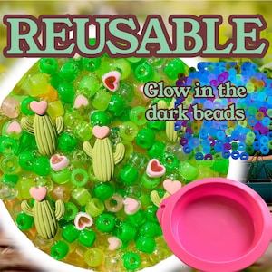 Op de afbeelding: Een verzameling groene kralen, cactusvormige bedels en hartvormige bedels. De afbeelding bevat ook een roze kom en een stapel blauwe en groene glow-in-the-dark kralen. Het woord "REUSABLE" staat bovenaan de afbeelding.