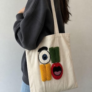 Op de afbeelding: Crèmekleurige draagtas met een grillig ontwerp. Het ontwerp bevat een groot oog, groene en gele rechthoeken en een rode mond. De tas heeft een eenvoudige, rechthoekige vorm met een lange riem.
