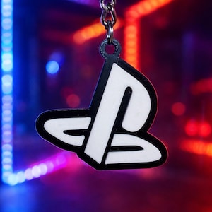 May include: A black and white PlayStation logo keyring. The keyring features the iconic PlayStation symbol, with a white design outlined in black. The background has a blurred, colourful neon light effect.
