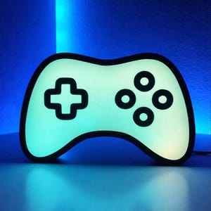 Könnte beinhalten: Eine leuchtende, hellgrüne Controller-förmige Lampe mit schwarzem Umriss. Die Lampe hat ein Steuerkreuz und vier kreisförmige Knöpfe. Der Hintergrund ist ein Farbverlauf aus Blautönen, der eine lebendige und moderne Ästhetik erzeugt.