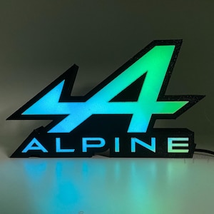 Könnte beinhalten: Ein schwarz-grünes beleuchtetes Schild mit dem Wort "ALPINE" und einem stilisierten "A"-Logo. Das Schild hat einen Farbverlauf, der von Blau nach Grün übergeht. Das Schild ist beleuchtet und eingesteckt.