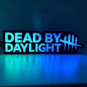 Pode incluir: Uma placa retangular preta com as palavras "DEAD BY DAYLIGHT" em verde-azulado brilhante e branco. A placa também apresenta um gráfico de uma mão com quatro linhas verticais riscadas. A placa é iluminada com luz verde-azulado e azul.