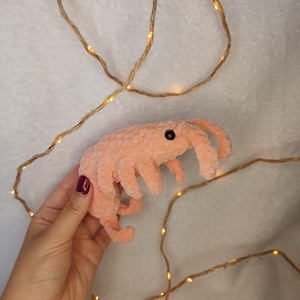 May include: A pink crocheted plush toy shaped like a sea creature with black eyes. The toy is being held in a hand against a background of string lights.