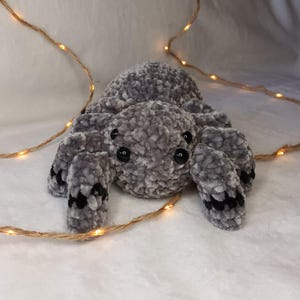 Puede incluir: Un juguete de peluche de araña de crochet gris con ojos negros y detalles negros en las patas.
