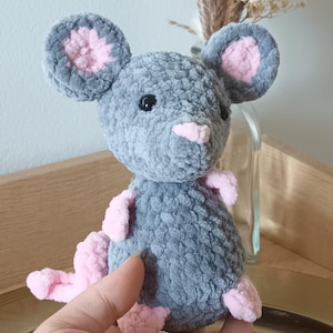 Peut inclure: Un jouet en peluche de souris gris crocheté avec des oreilles et des pieds roses. La souris a des yeux noirs et un nez rose.