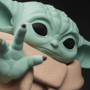 May include: A close-up of a light green figurine of a character with large black eyes and pointed ears. The figurine is reaching out with a hand, and is wearing a tan-colored garment. The figurine is a character from a popular science fiction franchise.