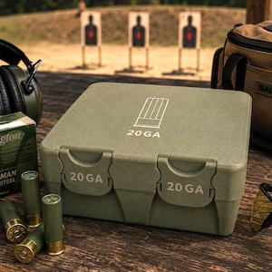 May include: An olive green plastic ammunition storage box with the text 20 GA printed on the lid and two latches. The box is surrounded by a box of Remington 20 gauge shotgun shells, four loose green shotgun shells, black ear protection, and glasses.