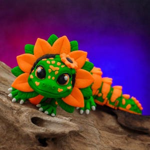 May include: A green and orange lizard figurine featuring a large sunflower-shaped collar around its head. The lizard has large black eyes, small white claws, and a segmented body with orange spots and patterns, resembling a floral creature.
