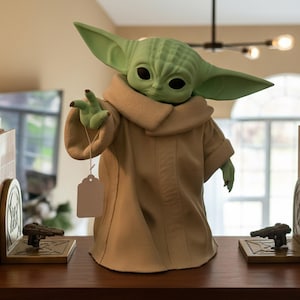 May include: A green figurine of a character from a popular science fiction franchise, wearing a tan robe and holding a blank tag. The figurine has large ears and black eyes. Two bookends with gun-shaped designs flank the figurine.