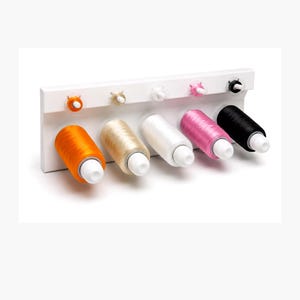 May include: A white wall-mounted thread rack holding five spools of sewing thread in orange, beige, white, pink, and black. The rack features five horizontal pegs for the spools and five smaller pegs above each spool for additional thread storage.