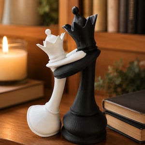 May include: A decorative sculpture featuring two chess pieces, a white queen and a black queen, positioned in an embrace. The white queen is slightly smaller and leans into the taller black queen, with both figures displaying detailed crowns.