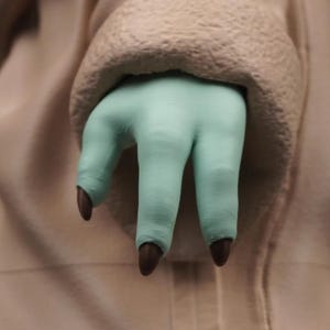 May include: A close-up of a light green hand with three fingers and dark brown nails. The hand is emerging from a beige, textured fabric. The fingers are slightly curved, and the nails are pointed.