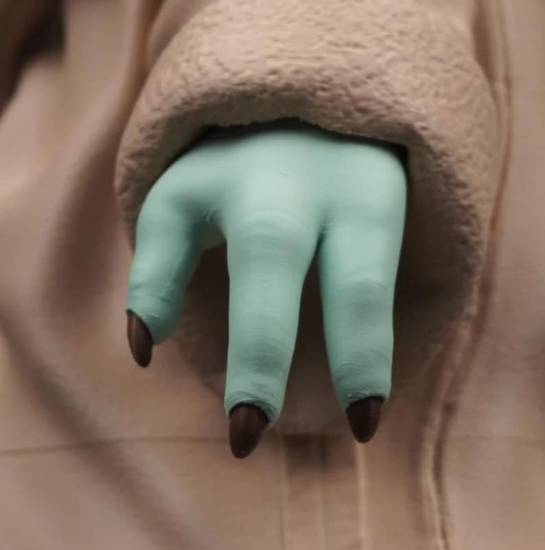 May include: A close-up of a light green hand with three fingers and dark brown nails. The hand is emerging from a beige, textured fabric. The fingers are slightly curved, and the nails are pointed.