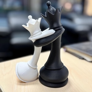 May include: A black and white chess piece sculpture showing two chess queens embracing. The white queen leans into the black queen, with their crowns touching. The pieces are on a light wood surface.
