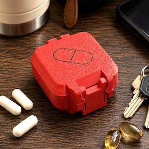 May include: A square, red, 3D-printed pill organizer box with a hinged lid featuring a black outline of a capsule icon. The container has a textured surface and is surrounded by two white cylindrical capsules and two translucent yellow gel capsules.