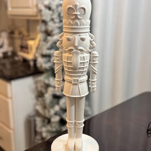 May include: A cream-colored, decorative nutcracker figurine. The figurine has a detailed design, including a crown, mustache, and uniform. It stands on a round base and is approximately 12 inches tall, a classic holiday decoration.