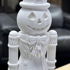 May include: A white, 3D-printed Halloween-themed figurine. The figure is a jack-o'-lantern wearing a top hat and a decorative jacket with a bow tie. The figurine has a smiling face and cylindrical arms.