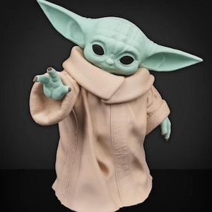 May include: A figurine of a character with large ears and large black eyes, wearing a tan robe with a cream-colored collar. The character's skin is a light green color, and it is reaching out with one hand.