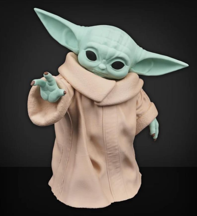 May include: A figurine of a character with large ears and large black eyes, wearing a tan robe with a cream-colored collar. The character's skin is a light green color, and it is reaching out with one hand.