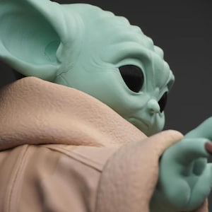 May include: A close-up of a figurine of a character with large ears, black eyes, and a light green complexion. The character is wearing a tan-colored robe. The figurine is a collectible item.