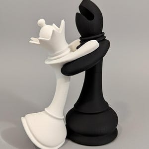 May include: A black and white chess set sculpture. The white queen is embraced by the black bishop. The queen has a crown and the bishop has a tall, open head. The chess pieces are on a gray background.