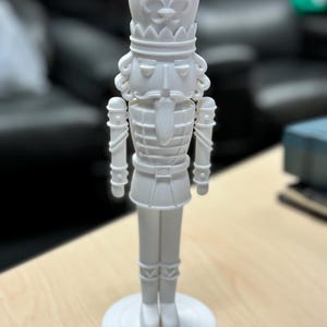 May include: A white, detailed nutcracker figurine. The figurine has a crown, a stern face, and a uniform with decorative elements. It stands on a circular base. The background is blurred, suggesting an indoor setting.