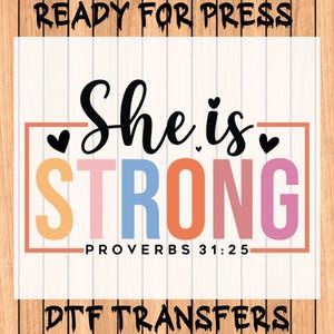 May include: A DTF transfer design with the words "She is STRONG" in black script and colorful block letters. The design includes the Bible verse Proverbs 31:25 and is framed in a peach-colored rectangle. The words "READY FOR PRESS" and "DTF TRANSFERS" are also included.