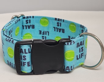 May include: A blue dog collar with a white tennis ball pattern and the text "Ball is Life". The collar has a black buckle and a silver D-ring.
