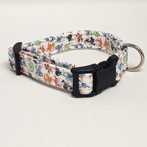 May include: A white dog collar with a colorful dragon print and a black plastic buckle. The collar has a silver metal D-ring.