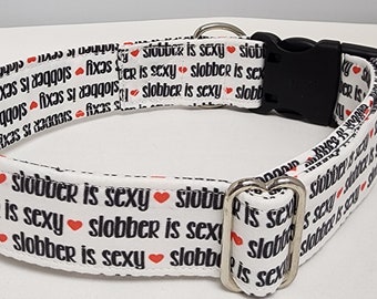 May include: White dog collar with a black buckle and silver D-ring. The collar has a repeating pattern of the text "Slobber is Sexy" with a red heart between each phrase.