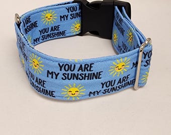 May include: A light blue dog collar with a black buckle and silver D-ring. The collar has a repeating pattern of yellow suns with smiling faces and the text "You are my sunshine".