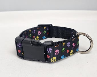 May include: Black dog collar with a colorful paw print pattern. The collar has a black buckle and a silver D-ring.