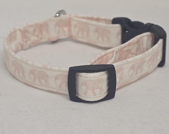 May include: A light pink and white patterned pet collar featuring a repeating elephant design. The collar has a black plastic buckle and a silver metal loop for attaching a leash. The collar is designed for small to medium sized pets.