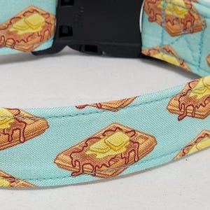 May include: A dog collar with a repeating pattern of waffles with butter on a light blue background. The collar has a black plastic buckle.