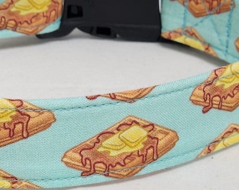 Puede incluir: Un collar para perros con un patrón repetido de gofres con mantequilla sobre un fondo azul claro. El collar tiene una hebilla de plástico negro.