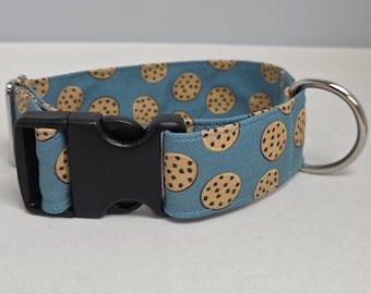 May include: A blue dog collar with a repeating pattern of brown cookies. The collar has a black plastic buckle and a silver metal D-ring for leash attachment. The collar is designed for pets.