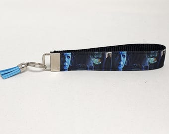 May include: A black and blue key chain with a blue tassel and a silver key ring. The key chain features a repeating pattern of a man's face.