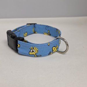 Puede incluir: Un collar para perros azul claro con una hebilla de plástico negro y una anilla en D de metal plateado. El collar está estampado con personajes de dibujos animados amarillos.
