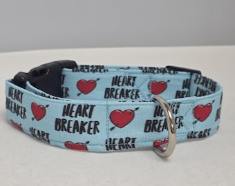 May include: A light blue pet collar featuring a repeating pattern of red hearts pierced by arrows and the black text "HEART BREAKER". The collar includes a black plastic side-release buckle and a silver-toned metal D-ring for attaching a leash.